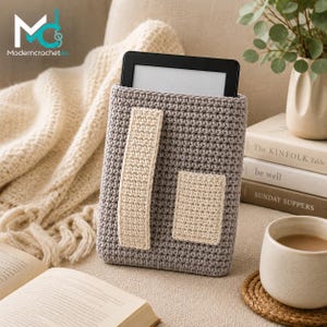 Peut inclure: Une housse au crochet grise pour liseuse électronique, dotée d'une sangle verticale couleur crème et d'une petite poche rectangulaire couleur crème sur le devant. La housse présente un motif de mailles dense et uniforme et contient une liseuse noire.