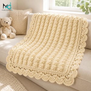 May include: A cream-colored crochet blanket featuring a textured vertical bobble stitch pattern and a decorative scalloped border. The blanket is draped over a sofa next to a plush teddy bear with a ribbon bow around its neck.