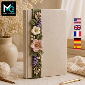 May include: A rectangular olive green crochet bookmark featuring a vertical arrangement of three-dimensional flowers and leaves in shades of pink, cream, and purple. The bookmark is placed vertically against the spine of a light beige hardcover book.