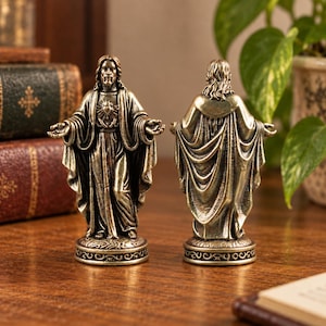 Peut inclure: Deux figurines métalliques aux tons bronze représentant Jésus-Christ sur des socles circulaires. Une figure fait face avec les bras tendus, montrant un emblème du Sacré-Cœur sur la poitrine, tandis que l'autre est vue de dos avec des robes drapées.