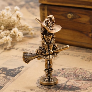 Puede incluir: Una pequeña figura de metal color bronce de un personaje con un sombrero de ala ancha con un lazo, de pie sobre una base circular. La figura sostiene una escoba horizontalmente y lleva botas, una falda y una parte superior detalladas.