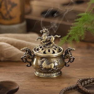 May include: A metallic bronze-toned incense burner featuring a round body with embossed horse figures on the sides and handles shaped like horses. The lid has a perforated design topped with a horse figurine, emitting thin wisps of smoke.