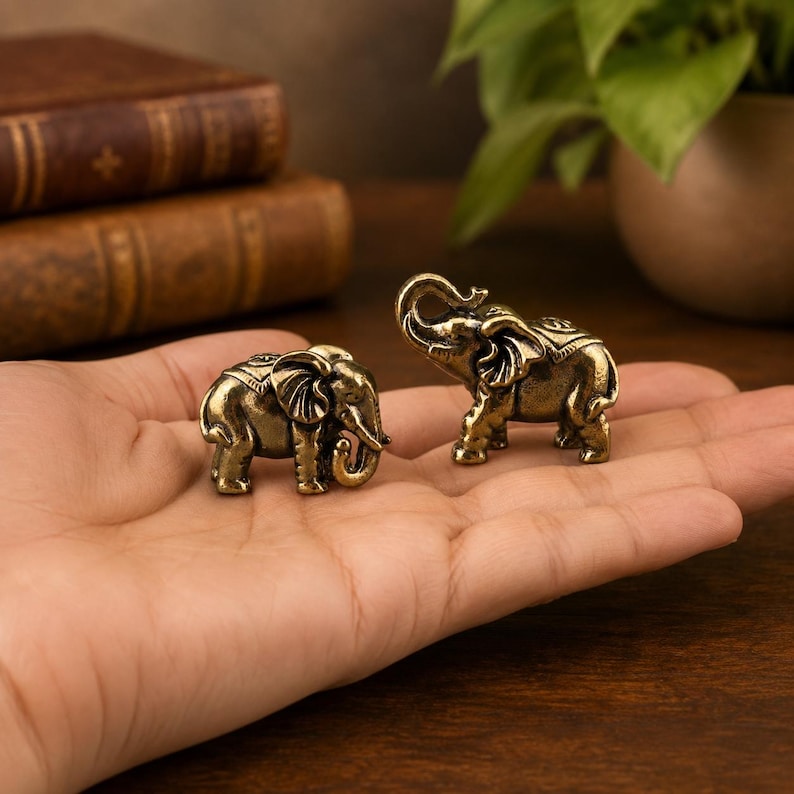 May include: Two small, antique-style brass elephant figurines with detailed textures. One elephant stands with its trunk curled downward, while the other stands with its trunk raised upward. Both figures feature decorative saddle patterns on their backs.