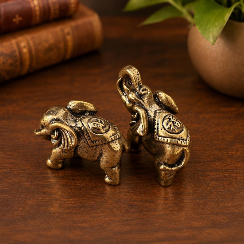 May include: Two small brass elephant figurines with a weathered, antique finish. Each elephant features a decorative saddle blanket on its back, engraved with a circular Chinese character. One elephant stands upright with its trunk raised, while the other is positioned in a walking stance.