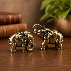 May include: Two small, metallic gold-toned elephant figurines. One elephant stands with its trunk curled downward, while the other stands with its trunk raised upward. Both figures feature detailed textures, including etched patterns on their backs.