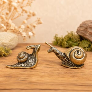May include: Two small brass snail figurines with textured, bumpy skin and smooth, spiraled shells. One snail is positioned with its head raised, while the other is depicted in a crawling posture with its head extended forward on a wooden surface.