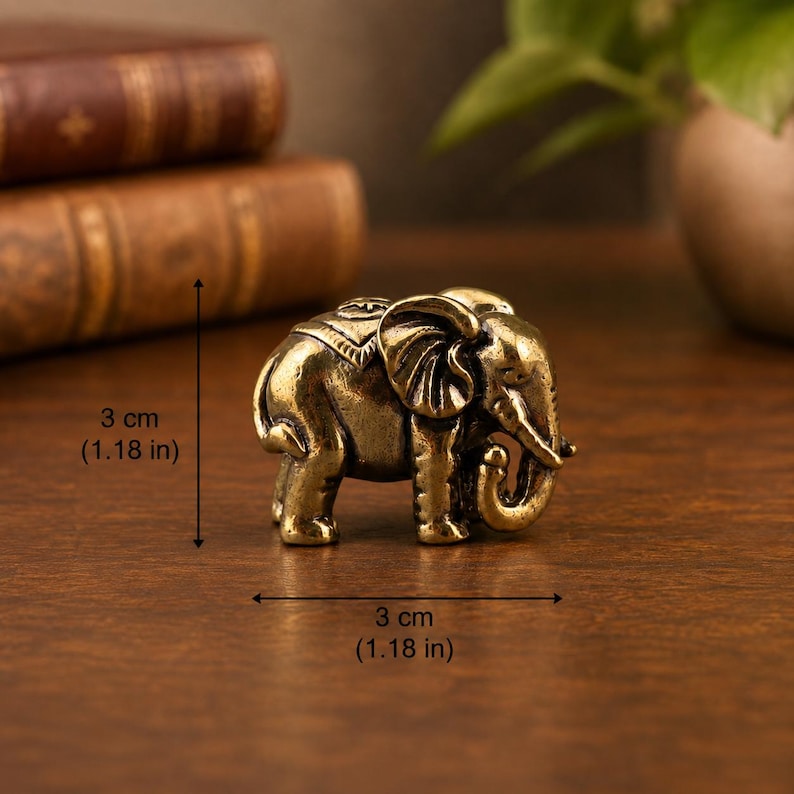 May include: A small brass elephant figurine with an ornate saddle design. The figure measures 1.18 inches in height and 1.18 inches in length, featuring a trunk curled downward and textured ears, standing in a profile orientation on a flat surface.
