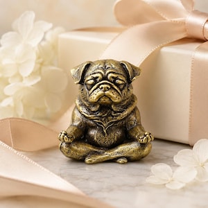 May include: A small, antique gold-toned figurine of a pug dog sitting in a cross-legged meditation pose with its eyes closed and paws resting on its knees. The sculpture features detailed facial wrinkles and a textured, weathered metallic finish.