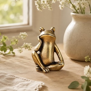 Puede incluir: Una pequeña figura de rana de metal en tono dorado, representada en una pose sentada y pensativa. La rana tiene una cabeza lisa y redondeada, ojos prominentes y una mano apoyada en la mejilla. La superficie tiene un acabado texturizado.