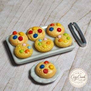 May include: A miniature rectangular white tray holding six small round cookies topped with colorful candy pieces, accompanied by a single cookie on a small scalloped white plate and a pair of silver-toned metal tongs beside the tray.
