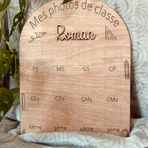 Può includere: Una tavola in legno ad arco per foto scolastiche con il testo inciso Mes photos de classe e il nome Roman. Include dodici spazi rettangolari etichettati con i livelli scolastici francesi da PS a 3ème, decorati con icone scolastiche incise.