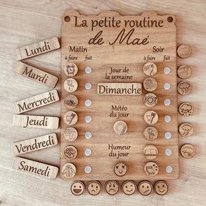 Könnte beinhalten: Ein hölzernes Tagesablauf-Board mit der Aufschrift La petite routine de Mae und Spalten für Morgen- und Abendaufgaben. Es enthält abnehmbare Holzplättchen für Wochentage, Wettersymbole und Stimmungsgesichter mit eingravierten Texten.