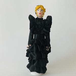May include: A small figurine with blonde hair, blue eyes, and red lips, wearing a black dress with long sleeves, a high collar, and multiple layers of ruffled fabric. The dress features a wide black waistband and a textured bodice with buttons.