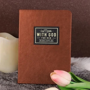 May include: A brown, rectangular, leather-textured journal with a black rectangular label on the front cover. The label features the text "One Minute WITH GOD FOR MEN 365 DAILY DEVOTIONS" printed in white and gold lettering with a thin gold border.