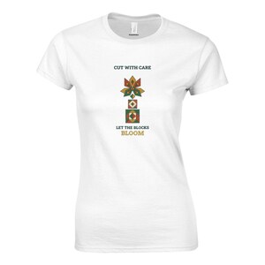 Può includere: Una t-shirt bianca a maniche corte con un design grafico che riporta il testo CUT WITH CARE e LET THE BLOCKS BLOOM. Il disegno include una disposizione verticale di forme geometriche nei toni del verde, rosso e giallo, simile a un fiore.