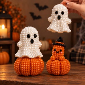 Op de afbeelding: Drie decoratieve figuren gemaakt van kleine kraaltjes: een witte geest met zwarte ogen en mond, een tweede witte geest zittend op een oranje pompoen, en een oranje pompoenfiguur met een zwarte hoed, een lachend gezicht en roze wangen.