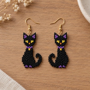 May include: A pair of drop earrings featuring a black cat design crafted from small beads. The cats have yellow eyes, orange noses, and purple collars, suspended from gold-toned metal hooks. The beadwork creates a structured, pixelated cat silhouette.