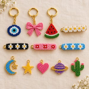 May include: A collection of beaded accessories including three gold-toned keychains with charms shaped like an evil eye, a pink bow, and a watermelon slice. Three rectangular hair clips feature daisy patterns in white, black, and blue. Five charms include a blue crescent moon with a star, a yellow star, a pink heart, a purple planet, and a green cactus.