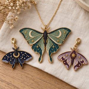 Può includere: Una collana a catena dorata con un grande ciondolo a forma di falena in perline verdi e verde acqua con motivi a mezzaluna e stelle, accompagnata da due ciondoli a forma di falena più piccoli in blu scuro e viola con motivi celesti e chiusure a moschettone.