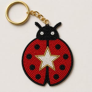 May include: A beaded ladybug keychain featuring a red body with black spots and a central white star outlined in gold beads. The ladybug has a black head with two small white flower-like accents and is attached to a gold-toned metal split ring.