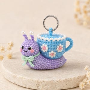 May include: A beaded snail figurine featuring a purple body with a light green bow and a blue teacup shell decorated with white and pink floral patterns. The snail has two antennae topped with white beads and a silver metal ring on top.