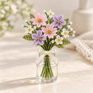 Puede incluir: Una pequeña botella de vidrio que contiene un ramo de flores de cuentas hechas a mano en tonos rosa, morado y blanco con hojas de cuentas verdes. Los tallos están atados con un pequeño lazo de tela color crema en el cuello de la botella.
