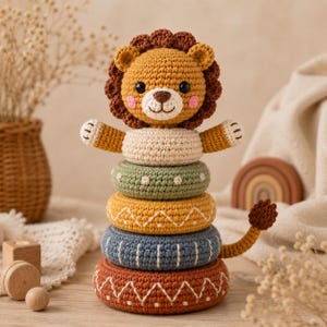 May include: A crochet lion stacking toy featuring a golden-brown lion head topper with a dark brown mane and pink cheeks. The toy consists of four stacked rings in sage green, mustard yellow, slate blue, and terracotta, with a small lion tail.