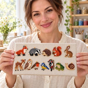 May include: A collection of small, beaded animal charms arranged on a rectangular beige display board. The charms include a fox, badger, hedgehog, squirrel, rabbit, deer, owl, robin, blue tit, woodpecker, frog, and snail, each with a gold loop.