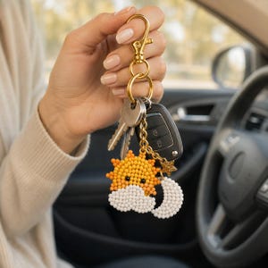 May include: A gold-toned metal keychain featuring a beaded sun, a white beaded cloud, and a white beaded crescent moon charm. The keychain includes a gold lobster clasp, a split ring, and a small gold star charm attached to a black car key fob.