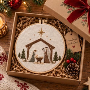 May include: A round wooden embroidery hoop containing a cream-coloured fabric with a stitched nativity scene featuring a stable, a star, and figures. A brown gift box includes a tag with the text Handmade with Love and a pinecone decoration.