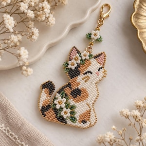 Könnte beinhalten: Ein Glückskatzen-Schlüsselanhänger aus kleinen Perlen in Weiß, Beige und Schwarz. Die Katze trägt einen Blumenkranz und hat ein Bündel weißer Blüten mit grünen Blättern an der Seite. Er ist mit einem goldenen Karabinerhaken versehen.
