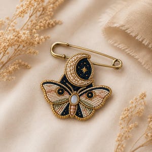 May include: A beaded brooch shaped like a moth with a crescent moon above it, attached to a gold-toned safety pin. The moth features intricate beadwork in shades of navy blue, cream, and soft pink, with a central oval stone and a gold star detail.