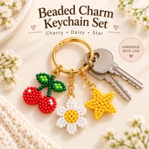 May include: A gold-toned metal keychain featuring three beaded charms: a red cherry with green leaves, a white daisy with a yellow centre, and a yellow star. The text reads Beaded Charm Keychain Set, Cherry, Daisy, Star, and Handmade with Love.