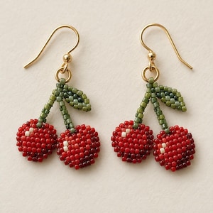 May include: A pair of drop earrings featuring two cherries crafted from small red, dark red, and white beads. The stems and leaves are made of olive green beads. Each earring hangs from a gold-toned metal hook wire.