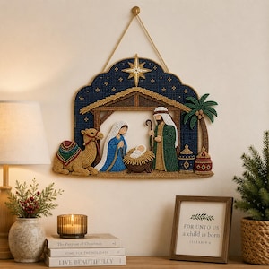 Może przedstawiać: Wisząca dekoracja wykonana techniką diamond painting przedstawiająca scenę narodzenia z wielbłądem, Marią, Józefem i dzieciątkiem Jezus w żłobie pod gwiazdą. Scena umieszczona jest w ciemnoniebieskiej, łukowatej ramie ze złotym sznurkiem do zawieszenia.