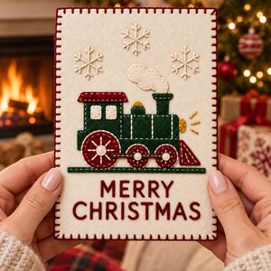 Puede incluir: Una tarjeta rectangular de fieltro color crema con una locomotora de vapor verde oscuro con ruedas y techo rojos. La tarjeta está decorada con copos de nieve bordados en blanco, una nube de humo y el texto bordado MERRY CHRISTMAS.