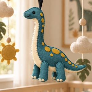 Puede incluir: Un adorno de dinosaurio de fieltro en color verde azulado y crema con manchas amarillas, con costuras visibles a lo largo de los bordes. El dinosaurio está suspendido por una cinta negra e incluye una pequeña cuenta negra como ojo y dedos de fieltro.