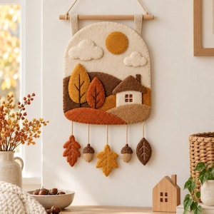 Op de afbeelding: Een vilten wandhanger met een herfstlandschap met een huis, bomen, wolken en een zon. Het stuk hangt aan een houten stok met touw en bevat bungelende vilten bladeren en eikels in tinten oranje, bruin en crème.