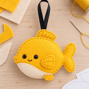 May include: A yellow felt fish ornament with a cream-colored belly, black bead eyes, and a stitched smile. The fish features a small side fin, a top fin, and a tail, all joined with visible blanket stitching. A black ribbon loop is attached to the top.