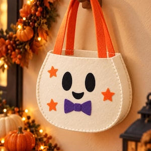 Puede incluir: Un bolso tipo cubo de fieltro blanco con dos asas naranjas, que presenta una cara de fantasma negra, una pajarita morada y tres apliques de estrellas naranjas. El bolso tiene costuras blancas visibles a lo largo de los bordes.