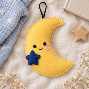 Peut inclure: Une décoration en forme de croissant de lune en feutrine jaune avec un bord cousu, deux yeux noirs circulaires, un petit sourire brodé, une joue rose circulaire et une petite étoile en feutrine bleu foncé fixée, suspendue par une boucle noire.