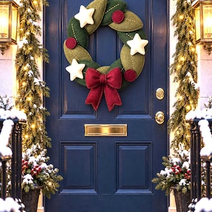 May include: A circular wreath constructed from layered felt leaves in shades of olive and forest green, accented with three cream-colored felt stars, three deep red felt circles, and a large, textured deep red felt bow at the bottom center.