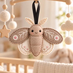 May include: A felt moth ornament in shades of beige and cream, featuring embroidered leaf patterns on the wings, small pink circular accents, and a black ribbon loop for hanging. The moth has a stitched face with two dark eyes and a smile.