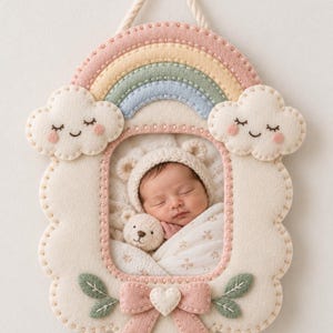 Op de afbeelding: Een vilten wandhanger met een pastelkleurige regenboogboog aan de bovenkant, twee glimlachende wolkjes aan de zijkanten en een roze strik met een hartje aan de onderkant. Het frame toont een foto van een slapende baby met een mutsje met oortjes.
