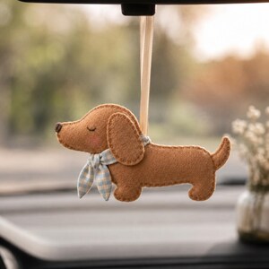 May include: A tan felt dachshund ornament hanging from a light beige ribbon. The dog has stitched closed eyes, a small dark brown nose, pink cheeks, and a light blue and white checkered fabric scarf tied around its neck with visible stitching.