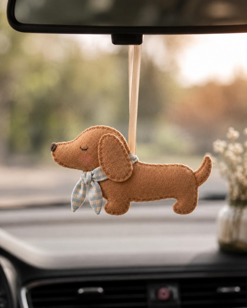 May include: A tan felt dachshund ornament hanging from a light beige ribbon. The dog has stitched closed eyes, a small dark brown nose, pink cheeks, and a light blue and white checkered fabric scarf tied around its neck with visible stitching.