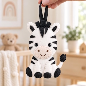 May include: A felt zebra ornament featuring white and black stripes, a light beige muzzle, and black stitched details. The zebra has two black eyes, a small stitched smile, and a black satin ribbon loop at the top for hanging purposes.