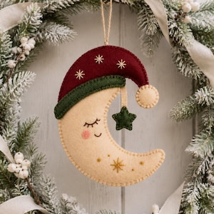 Pode incluir: Um ornamento de feltro em forma de lua crescente com um rosto sonolento, olhos fechados e bochechas rosadas. A lua usa uma touca de dormir vermelho-escura com uma borda verde-escura e uma pequena estrela verde-escura pendurada. Estrelas douradas bordadas decoram a lua.