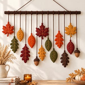 May include: A wall hanging featuring a horizontal dark brown rod suspended by a string. Hanging from the rod are various felt leaves in shades of red, orange, yellow, green, and brown, along with two felt acorns, all attached by brown cords.