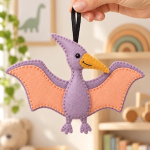 Peut inclure: Une décoration en feutrine en forme de ptérodactyle faite à la main, avec un corps et des ailes violettes dotés de panneaux de couleur pêche. Le dinosaure possède un bec jaune avec un sourire brodé, un petit œil noir et une boucle en ruban noir.
